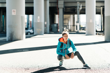 Portrait of young sporty woman sitting and resting under the bridge after workout in industrial city