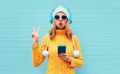 Winter portrait cool woman with phone in headphones listening to music showing peace hand sign...
