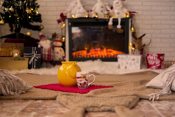 Teapot and cup of hot coffee, on the carpet surrounded by Christmas decorations and lights.