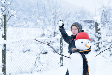   Winter teenager in a jacket and hat sculpts a snowman on a winter background with falling snow in frozen day concept