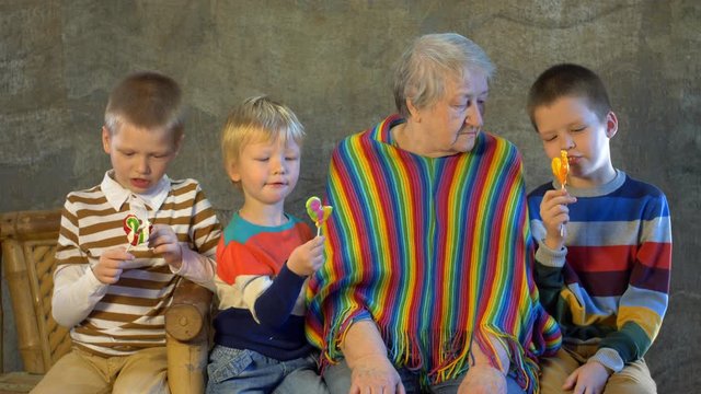 Three children in bright holiday dresses are holding caramel on stick. Children grouped around grandmother in anticipation of Christmas presents. Brothers love extravagant grandmother in bright poncho