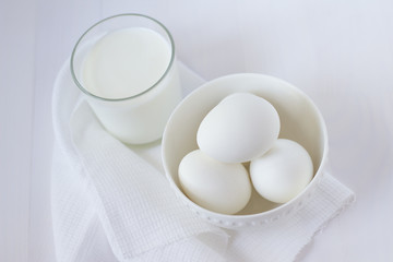 four white eggs lie in a plate on a white wooden table next to a towel and a glass of milk.