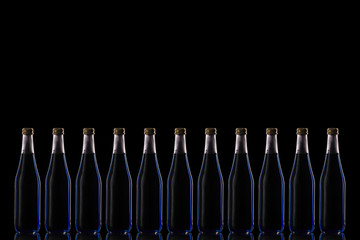 Eleven large glass bottles with a blue drink stand on a mirrored surface against a dark background in the center.