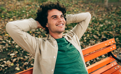 Good-looking young man with curly hair, smiling and looking up, feeling joyful, sitting on the...