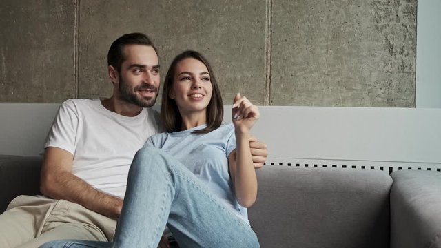 Cute cheerful young lovely couple hugging and speaking while looking at the window sitting on sofa at home