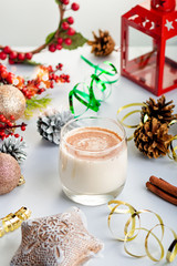 Traditional Christmas cocktail Eggnog with eggs, alcohol, grated nutmeg and cinnamon closeup. Sweet traditional drink on grey table with beige decorations, burning lanterns and pine cones