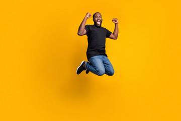 Fototapeta na wymiar Yeah i win. Full size photo of delighted funny crazy afro american guy celebrate lottery victory jump raise fists scream wear t-shirt denim jeans isolated bright color background