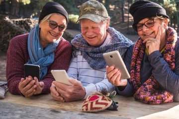 Front view of three retired people friends smiling and looking at the cellphones. Sitting outdoor in the forest. Wooden table. Pines in the background.