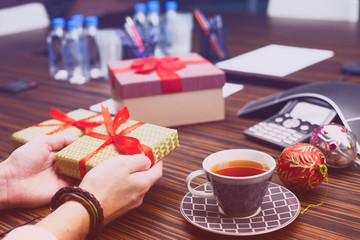 Young businessman surrounded by gifts in office. Workplace, cup of tea, laptop, ip phone and boxes. Christmas gift from colleague. Secret Santa office tradition. New year background.