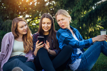 Three cute girls relax and socialize on the lawn in summer park. Young women sit on the green grass...