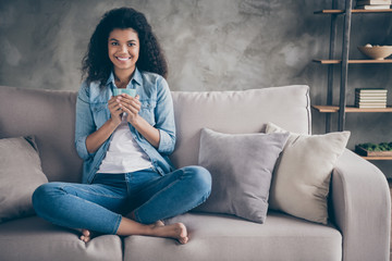 Obraz premium Photo of beautiful dark skin wavy lady crossed legs holding hot coffee beverage cup enjoy weekend at home sitting cozy couch casual denim outfit living room indoors