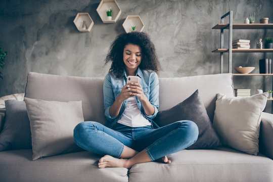 Photo of pretty dark skin curly lady homey domestic texting telephone with friends reading instagram comments sitting comfy couch casual denim outfit living room indoors