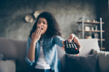 Photo of dark skin wavy lady homey mood holding tv remote control sleepy boredom yawning turn television off sitting comfy couch casual denim outfit flat indoors blurry focus