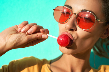 Close up portrait of a young stylish woman sexually licking a big red lollipop against blue background. - obrazy, fototapety, plakaty