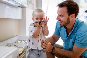 Health care, eyesight and vision concept. Little girl choosing glasses with father at optics store