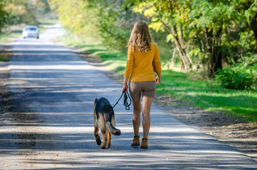 Naklejka na ściany i meble A happy girl walks away on a road in the woods with a German shepherd dog. The view from the back. A young teen puppy is a loyal friend guarding his mistress. On a yellow-green background of trees.