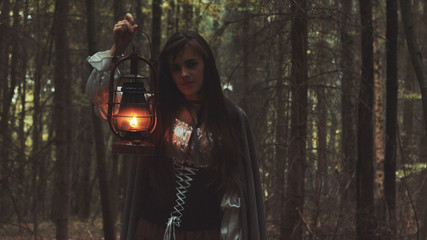 Naklejka premium Young girl with a lantern wandering old forest. Gothic scene from the movie.