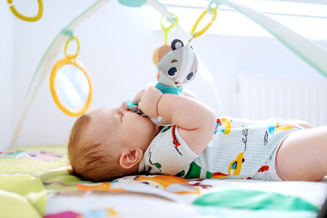 Adorable six months old boy lying in bed biting and playing with crib toys. Healthy upbringing concept.