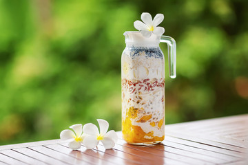 Jug of superfood layered smoothie with tropical fruits and berries at wooden table at green...
