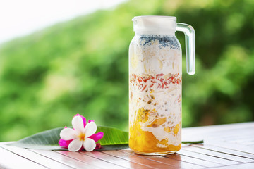 Pitcher with superfood layered smoothie with tropical fruits and berries at wooden table decorated with flowers at green tropical blurred background. Detox and healthy lifestyle.