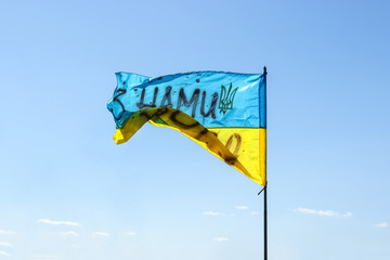 Waving Ukrainian yellow-blue flag against a blue sky. On the flag is the coat of arms of Ukraine and the inscription: 