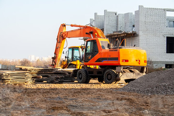 Modern orange excavator standing on the ground at a construction site next to wooden pallets and a white brick building under construction