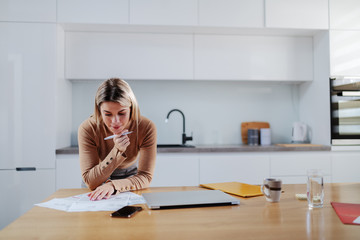 Cute caucasian blonde woman in sweater leaning on dining table and calculating bills. In hand is...