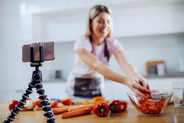 Attractive caucasian blonde woman in apron preparing vegetarian meal. In foreground is smart phone on tripod. Kitchen interior.