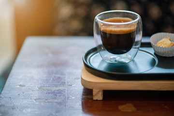 Hot espresso coffee in a clear glass, placed on a metal tray and wooden layer beside the coffee cup...