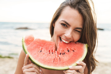 Image of young happy woman eating watermelon and winking