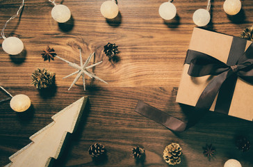 Present paper box wrapped with brown ribbon on wooden background with garland, decorative christmas tree and pine cones. Celebration and holiday concept