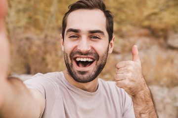 Image of unshaven laughing man showing thumb up and taking selfie photo