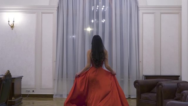 A girl in a red dress in a photo studio looks like a 19th-century Russian palace with high ceilings, she opens the door and goes to the window, moving slowly. She has a smile on her face.