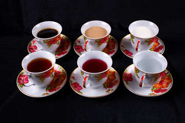 Six cups and saucers filled with water, juice, tea, coffee, milk and coffee with milk on a dark fabric background