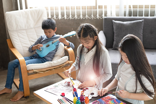 Two sisters painting colorful water color on paper,with interested feeling,young boy playing ukulele on wooden chair,doing activity together,at living room