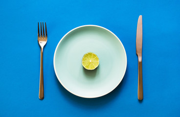 A slice of fresh lime lying on color plate with fork and knife on blue background.  Concept of vegan diet and citrus food.