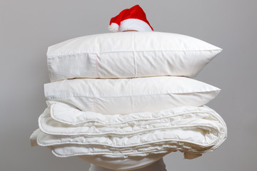 Woman in santa hat holding a warm duvet and feather pillows against a gray wall. Stack of bedding...