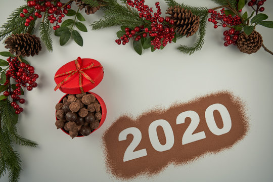 We welcome the year 2020 with a box of chocolates and chocolate truffles, some pineapples, some holly branches and cocoa powder on a white background.