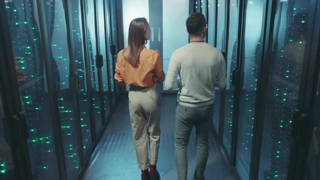Data center team walking in digital server room corridor. Back of man and woman colleagues talking examining rack server cabinets cooperating.