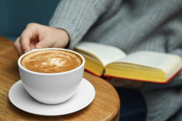 Woman reading book at table, focus on cup of coffee