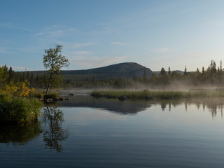 Beautiful morning over lake Sjabatjakjaure with haze mist in Sweden Lapland nature. Mountains, birch trees, spruce forest, rock boulders and grass. Sky, clouds and clear water.