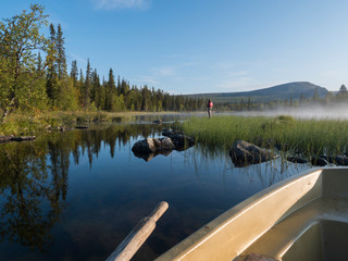 View from rowing boat on Fisherman man at lake Sjabatjakjaure in Beautiful sunny morning haze mist in Sweden Lapland nature. Mountains, birch trees, spruce forest. Blu sky background