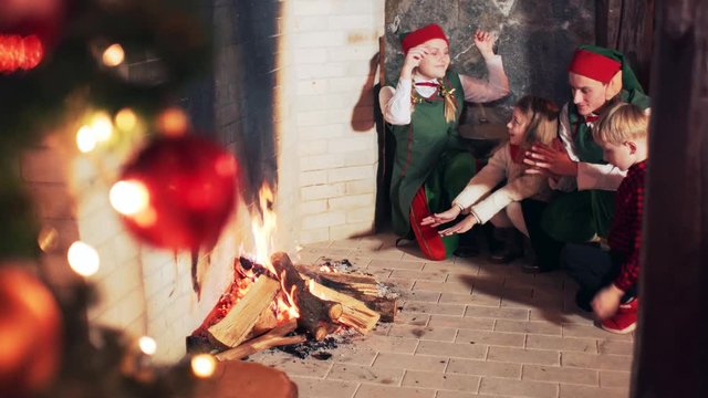 Happy children and Santa Claus elves warm their hands near the fireplace in the room with the Christmas tree.