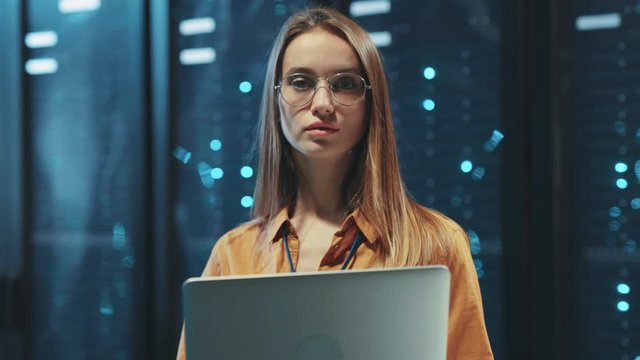 Female administrator working on laptop inspecting data center server room. Portrait of young woman IT specialist standing at row of rack server cabinets.