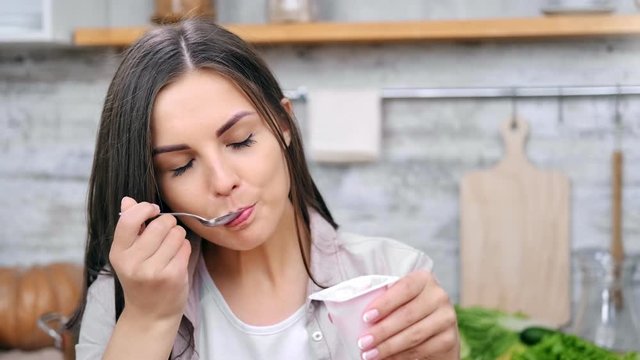 Close-up face of beauty young lady eating milky yoghurt using spoon posing looking at camera