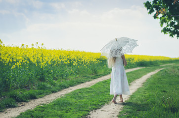 beautiful girl back view walking away with white sunshade umbrella along yellow rapeseed oil meadows