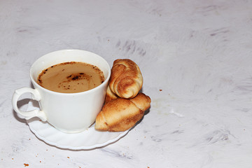 Cup of coffee with milk and fresh ruddy croissants on a gray table with breadcrumbs, place for text. Concept of a modern bakery. Delicious traditional breakfast.