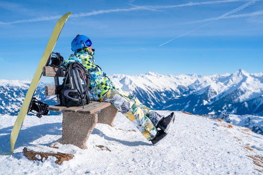 Image from side of athlete man sitting on bench next to snowboard in ski resort.