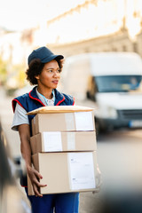 Female African American deliverer carrying cardboard boxes on the street.