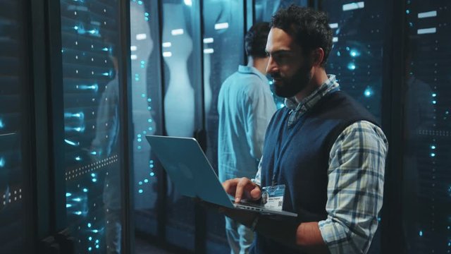 Bearded server specialist using computer laptop running diagnostics on server rack cabinets inspecting digital room of data center.